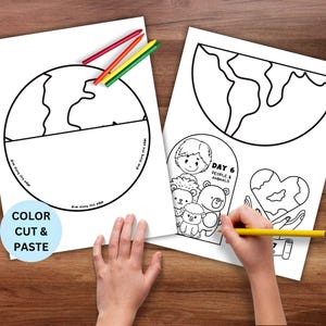 May include: Two white coloring pages with outlines of the Earth and illustrations of people and animals. The text "COLOR CUT & PASTE" is on one page. Crayons in red, yellow, and green are on the table. A hand is holding a yellow pencil.