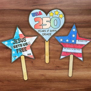 Può includere: Tre sagome di carta decorative su bastoncini di legno. Una sagoma a forma di cuore riporta la scritta USA 250 YEARS OF GOD'S GRACE. Una sagoma a stella riporta JESUS SETS US FREE 1776 2026. Un'altra stella presenta i colori della bandiera USA.