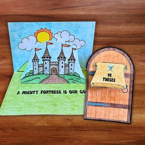 May include: A paper craft pop-up card featuring a gray castle with four towers under a yellow sun and blue sky, with the text A MIGHTY FORTRESS IS OUR GOD. Beside it, a wooden door cutout displays a scroll with a nail and the text 95 THESES.