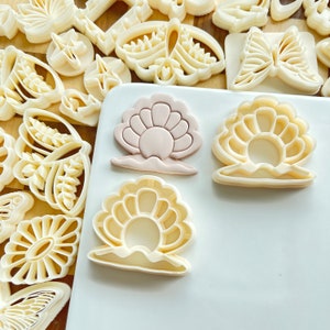 May include: Three cookie cutters shaped like seashells. The cutters are made of white plastic and have a detailed design. The seashells are arranged on a white surface.