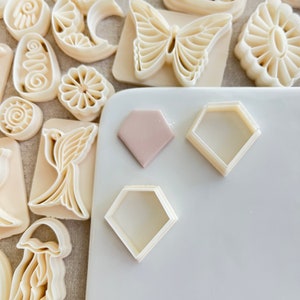 May include: A white surface with three cookie cutters, two are diamond shaped and one is a rounded rectangle. There is a light pink diamond shaped cookie cutter on the surface as well.