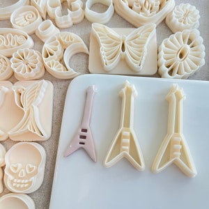 以下が含まれることがあります： 白いプラスチック製のクッキー型3つ。電気ギターの形をしています。クッキー型は白い表面の上に置かれています。