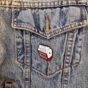 May include: A pin featuring a red and white cooler design with the text "DON'T HATE ME BECAUSE I'M A Little Cooler" is attached to a blue denim jacket. The pin has a white background with red accents and black text.