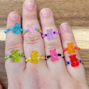 May include: A set of eight gummy bear rings, each with a different color and a beaded band. The rings are worn on a person's fingers.
