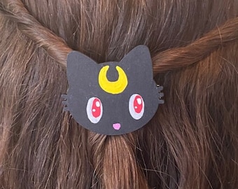 以下が含まれることがあります： 黄色い三日月とピンクの目をした黒い猫のヘアクリップ。