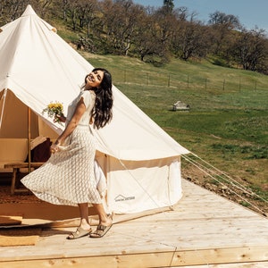 May include: A woman in a white crochet dress stands outside a white canvas tent with a bouquet of flowers in her hand. The tent is set up on a wooden deck with a grassy field and trees in the background.