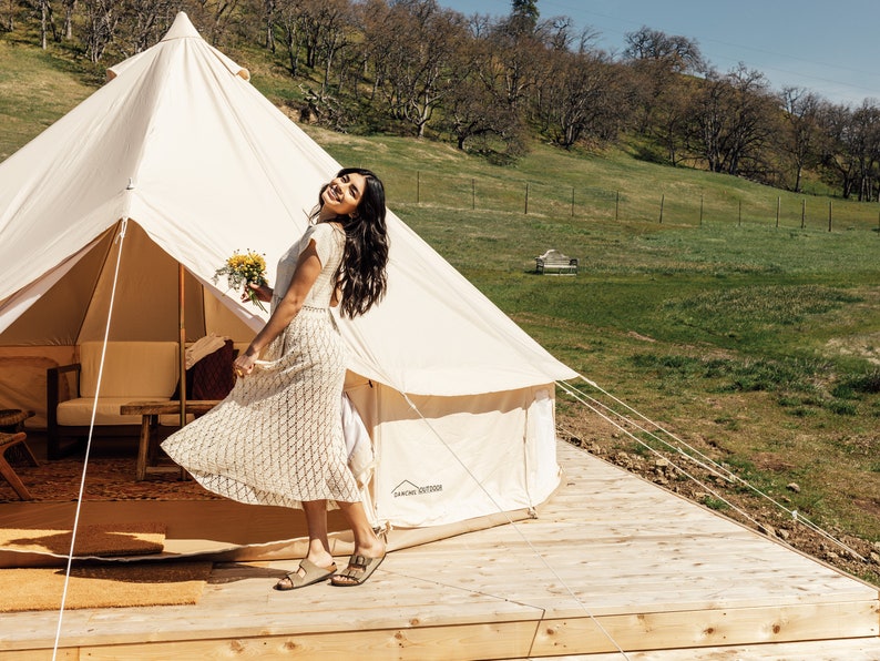 May include: A woman in a white crochet dress stands outside a white canvas tent with a bouquet of flowers in her hand. The tent is set up on a wooden deck with a grassy field and trees in the background.