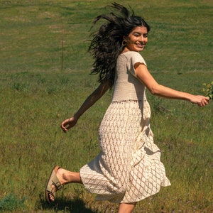 May include: A woman in a white crochet dress runs through a field of green grass, holding a bouquet of yellow flowers. She is smiling and looking over her shoulder.