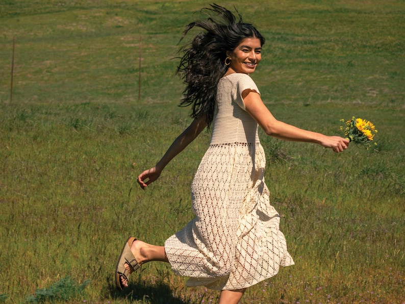May include: A woman in a white crochet dress runs through a field of green grass, holding a bouquet of yellow flowers. She is smiling and looking over her shoulder.