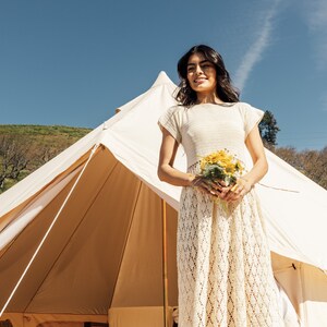 May include: A woman in a white crochet dress holds a bouquet of yellow flowers in front of a white tent.