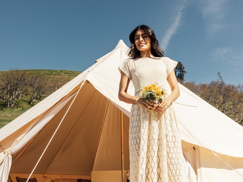 May include: A woman in a white crochet dress holds a bouquet of yellow flowers in front of a white tent.