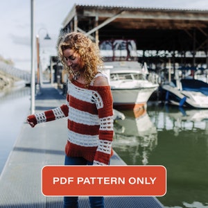 May include: A woman wearing a red and white striped crochet sweater with a loose, off-the-shoulder style. The sweater has a crocheted pattern and is worn over blue jeans. The woman is standing on a dock by the water. The text "PDF PATTERN ONLY" is on a red button.