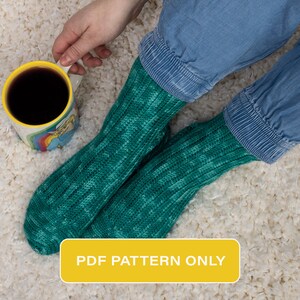 Puede incluir: Un patrón PDF para tejer calcetines azul verdoso con un patrón de punto texturizado. Los calcetines los lleva puestos una persona sentada en una alfombra blanca de pelo largo, sosteniendo una taza amarilla de café.