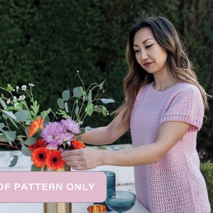 May include: A crochet pattern for a pink, open-stitch top with short sleeves. The pattern is available as a PDF download. The image shows a woman wearing the top and arranging flowers in a vase.