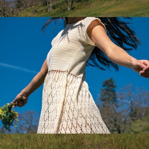 May include: A woman wearing a white crochet dress with a lacy pattern, standing in a field with a blue sky in the background.
