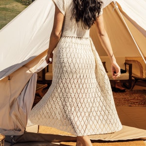 May include: A woman in a white crochet dress stands in the doorway of a white canvas tent. The tent has a wooden frame and is decorated with pillows and blankets.