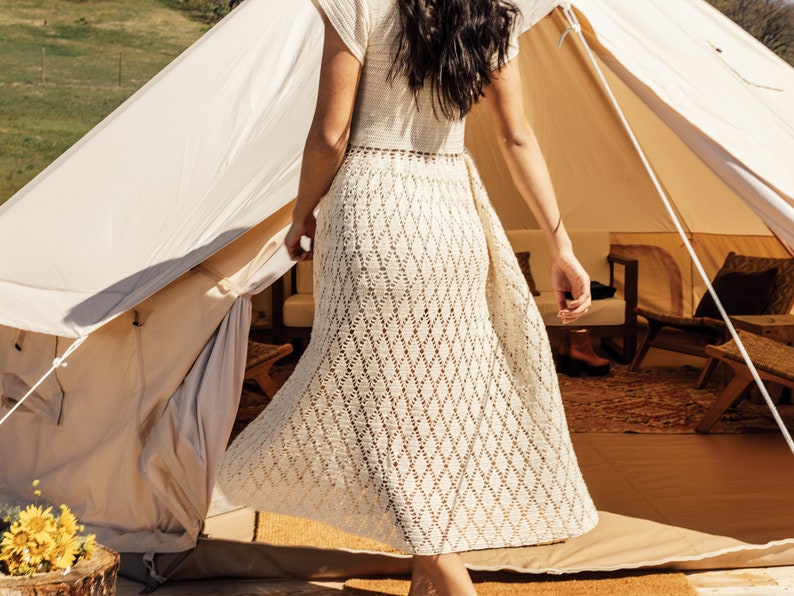 May include: A woman in a white crochet dress stands in the doorway of a white canvas tent. The tent has a wooden frame and is decorated with pillows and blankets.
