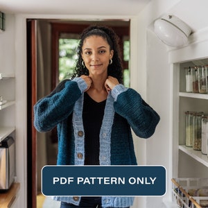 Könnte beinhalten: Eine Frau trägt eine blaue und hellblaue gehäkelte Strickjacke mit Knöpfen. Die Strickjacke ist nur ein PDF-Muster. Das Bild wurde in einer Küche mit Regalen voller Gläser aufgenommen.