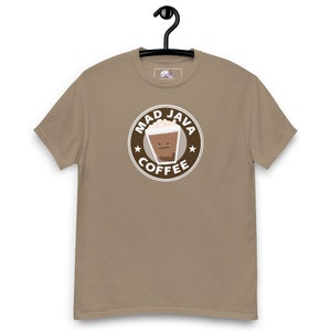 Può includere: Una maglietta color beige con un logo circolare marrone che dice "Mad Java Coffee" con una tazza di caffè di cartone animato con un viso al suo interno.