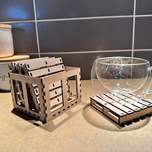 Op de afbeelding: Een set houten onderzetters in een houten kist, met een glazen beker op een enkele houten onderzetter. De onderzetters zijn gemaakt om eruit te zien als miniatuur pallets.