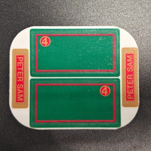 May include: An oval-shaped game piece with a white background. It features two green rectangles with red borders, each containing a red circle with the number 4. The text "PETER SAM" is printed vertically on gold rectangles.
