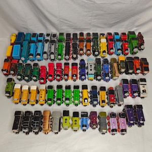 Puede incluir: Una colección de 50 locomotoras de tren de juguete de madera de colores. Las locomotoras están dispuestas en filas e incluyen varios personajes de la franquicia Thomas y sus amigos.