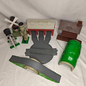 Könnte beinhalten: Ein Set mit Thomas der Lokomotive-Gleisabschnitten, darunter ein grüner Tunnel, eine graue Brücke, ein grauer Drehscheibe, ein rot-weißes Gebäude mit drei Garagentoren, ein braunes Gebäude mit einem Giebeldach, ein grauer Wasserturm und eine weiße Windmühle.