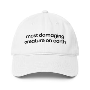 May include: White baseball cap featuring the black text "most damaging creature on earth" across the front. The cap has a curved peak and an adjustable fabric strap at the back.
