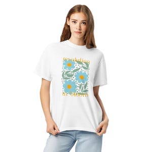 May include: White t-shirt with a floral design. It features light blue flowers, green leaves, and the text "revoluting in bloom" in yellow-gold. The shirt is made of a soft, comfortable material.