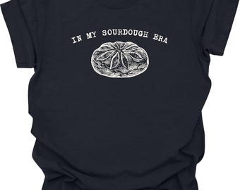 Könnte beinhalten: Ein marineblaues T-Shirt mit dem Text "IN MY SOURDOUGH ERA" über einer detaillierten weißen Illustration eines Brotlaibs. Das T-Shirt ist einfarbig und hat einen Rundhalsausschnitt.