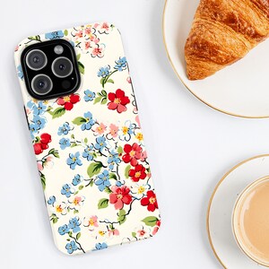 May include: A floral phone case featuring a cream background with a pattern of red, blue, and pink flowers and green leaves. The case has a camera cutout and is next to a plate with a croissant and a cup of coffee.