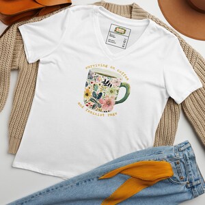 May include: White V-neck t-shirt with a floral coffee mug graphic and the text "surviving on coffee and feminist rage." The mug features pink, yellow, and blue flowers. The shirt is displayed with jeans and a tan sweater.