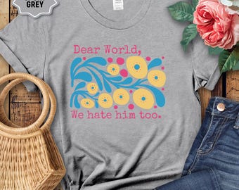 Op de afbeelding: Sportgrijs t-shirt met een bloemenontwerp in geel, blauw en roze. De tekst "Dear World, We hate him too." is in roze gedrukt. Het shirt wordt getoond met een geweven handtas en een spijkerbroek.
