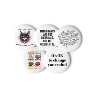 May include: A collection of five white round buttons with various text and graphic designs. One button features a black cat with text that reads "BORN TO DILLY DALLY" and "FORCED TO FIGHT FASCISM." Other buttons have phrases like "IMMIGRANTS ARE NOT CRIMINALS, BUT THE PRESIDENT IS."