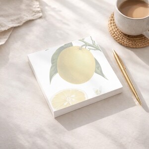 May include: A square notepad featuring a botanical illustration of a lemon with green leaves. A gold pen and a mug of coffee on a coaster are also visible. The notepad has a white background and a lemon slice illustration.
