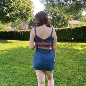 May include: A woman wearing a multi-colored crochet crop top with a denim skirt.