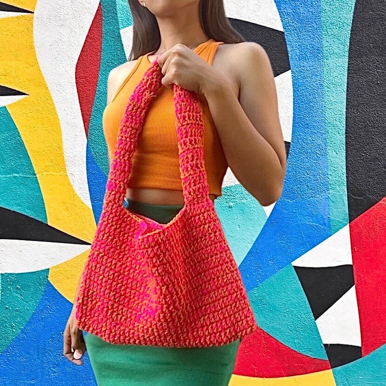 May include: A bright pink and orange crocheted tote bag with a long strap. The bag is being held by a person wearing a bright orange tank top and a green skirt.