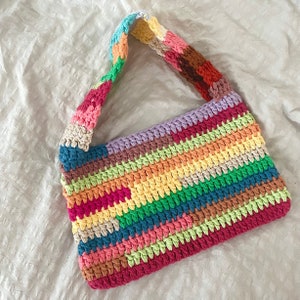 May include: A colorful crocheted shoulder bag with a rainbow striped pattern. The bag is made of yarn in shades of pink, red, orange, yellow, green, blue, and purple.