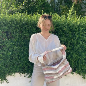 May include: A woman wearing a white, striped shirt holds a crocheted bag with pink, white, and brown stripes. The bag has a long strap and is made of thick yarn.