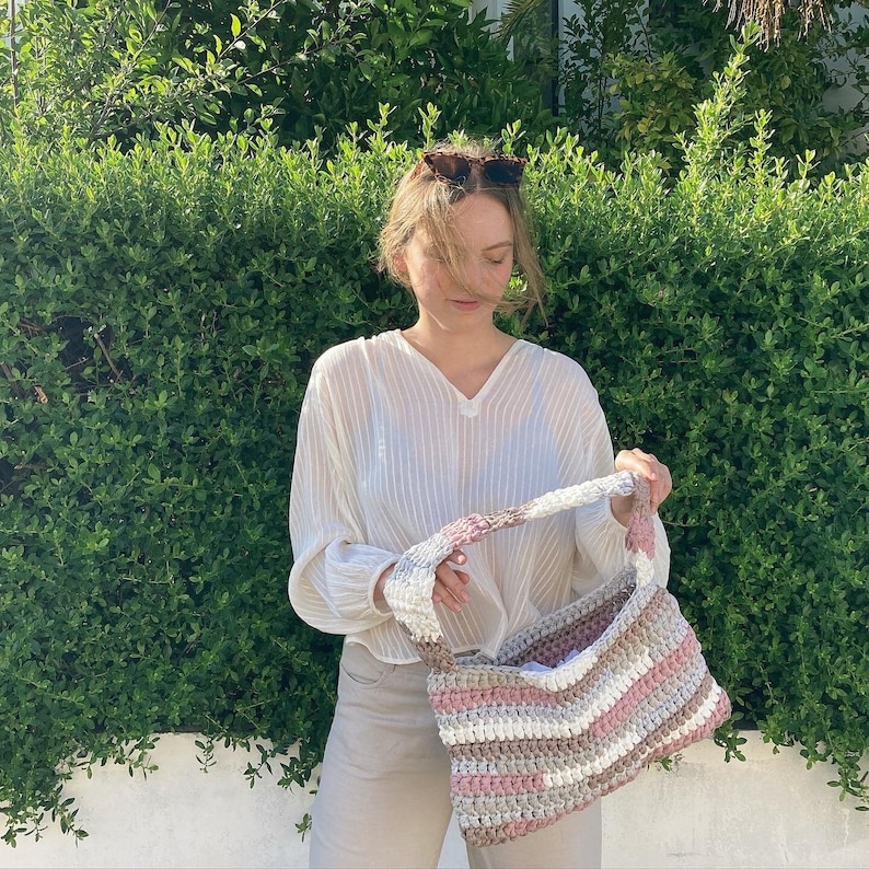 May include: A woman wearing a white, striped shirt holds a crocheted bag with pink, white, and brown stripes. The bag has a long strap and is made of thick yarn.