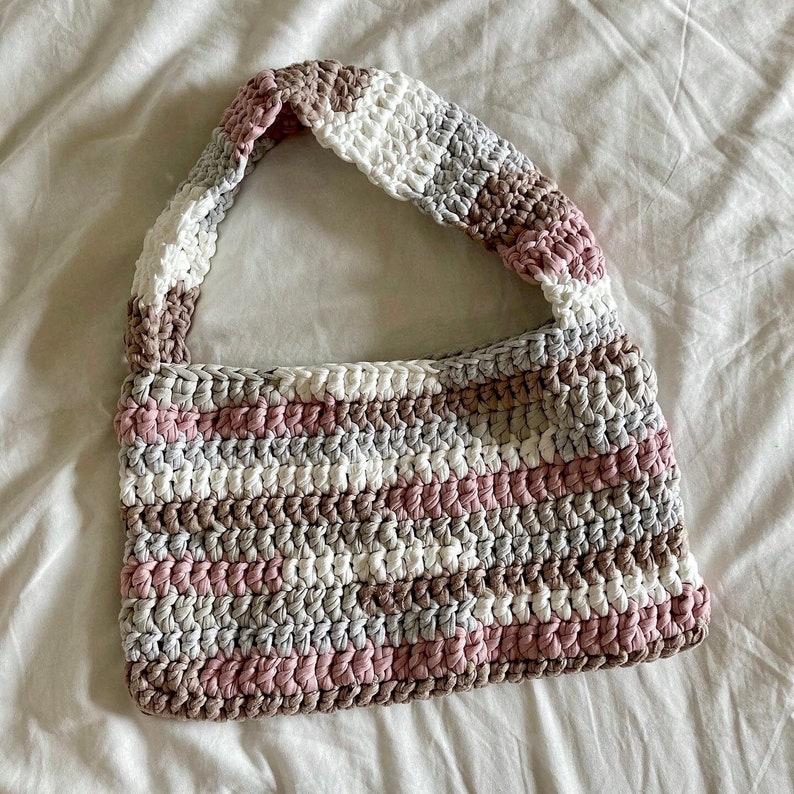 May include: A crocheted handbag with a pink, white, and brown striped pattern. The bag has a single strap and is made of a soft, textured yarn.