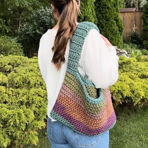 May include: A multi-colored crocheted tote bag with a teal, green, orange, and purple striped pattern. The bag is worn over the shoulder of a person wearing a white sweater and blue jeans.