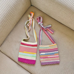 May include: Two colourful crocheted bags with striped patterns and long straps. The bag on the left is pink with light blue, yellow, and green stripes. The bag on the right is purple with blue, orange, and beige stripes.