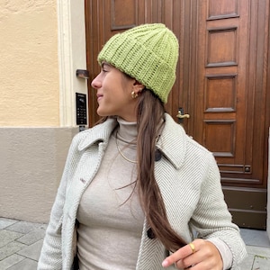 Könnte beinhalten: Eine Frau trägt eine hellgrüne Strickmütze mit Rippenstruktur. Sie trägt einen hellbeigen Rollkragenpullover und einen weiß-grauen Fischgrätenmantel.