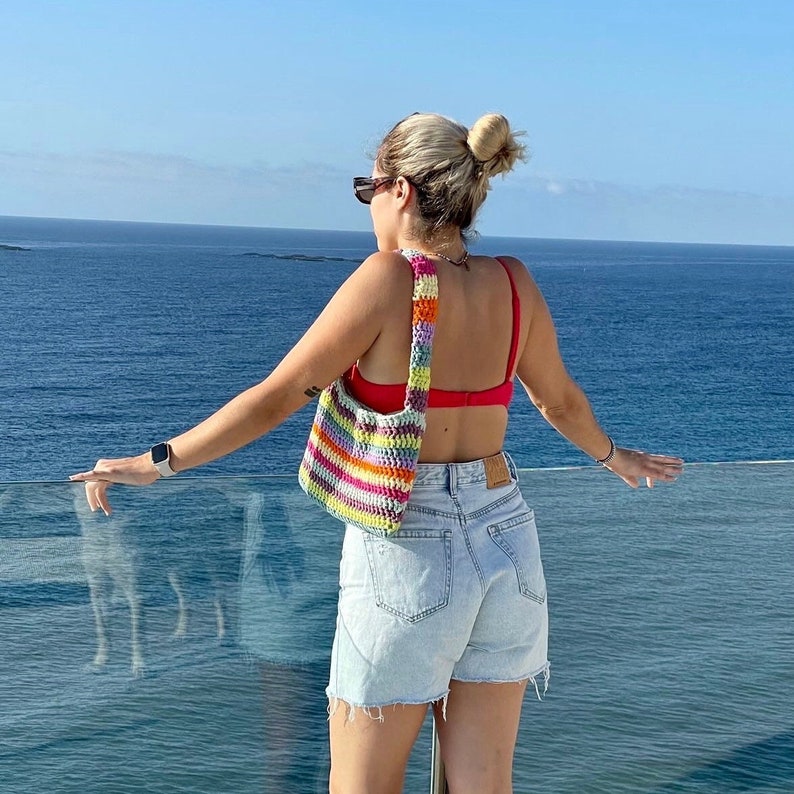 May include: A woman wearing a red bikini top and denim shorts stands on a balcony overlooking the ocean. She is carrying a colorful crocheted bag with a long strap.