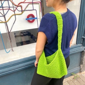 May include: A bright green crocheted shoulder bag with a long strap. The bag is worn by a person wearing a dark blue sweater. The bag has a unique, slouchy shape.