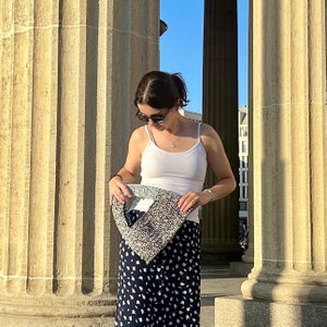 May include: A woman wearing a white tank top and a black skirt with white polka dots is holding a silver and gray crocheted bag. She is standing in front of a stone column.