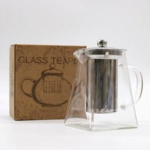 May include: A clear glass teapot with a stainless steel lid and infuser. The teapot has a square base and a handle on the right side. The teapot is sitting on a brown cardboard box with the words "GLASS TEAPOT" printed on it.