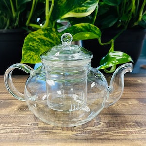 May include: A clear glass teapot with a built-in infuser. The teapot has a round body and a lid with a knob on top. The teapot is sitting on a wooden surface.