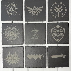 May include: Nine black square slate coasters with different engraved designs from the Legend of Zelda video game series. The designs include the Triforce, Master Sword, Hylian Shield, and more.